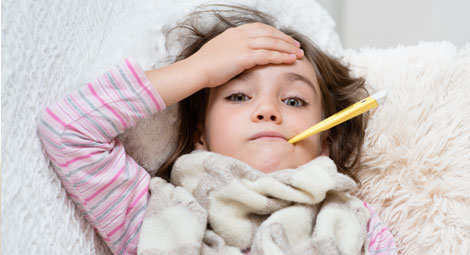 Sick child in a blue dressing gown with a thermometer in her mouth. She is holding her forehead.